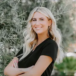 A woman with long, wavy blonde hair stands outside with arms crossed. She is wearing a black top and smiling. The background features greenery with blurred leaves.
